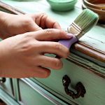**

"Close-up shot of a person's hands applying chalk paint to an ornate, antique wooden dresser. The dresser is partially painted in a distressed, pastel color (like a soft lavender or mint green). Paint brushes, sanding paper, and a jar of wax are visible in the background. Soft, natural light. French country style."

**